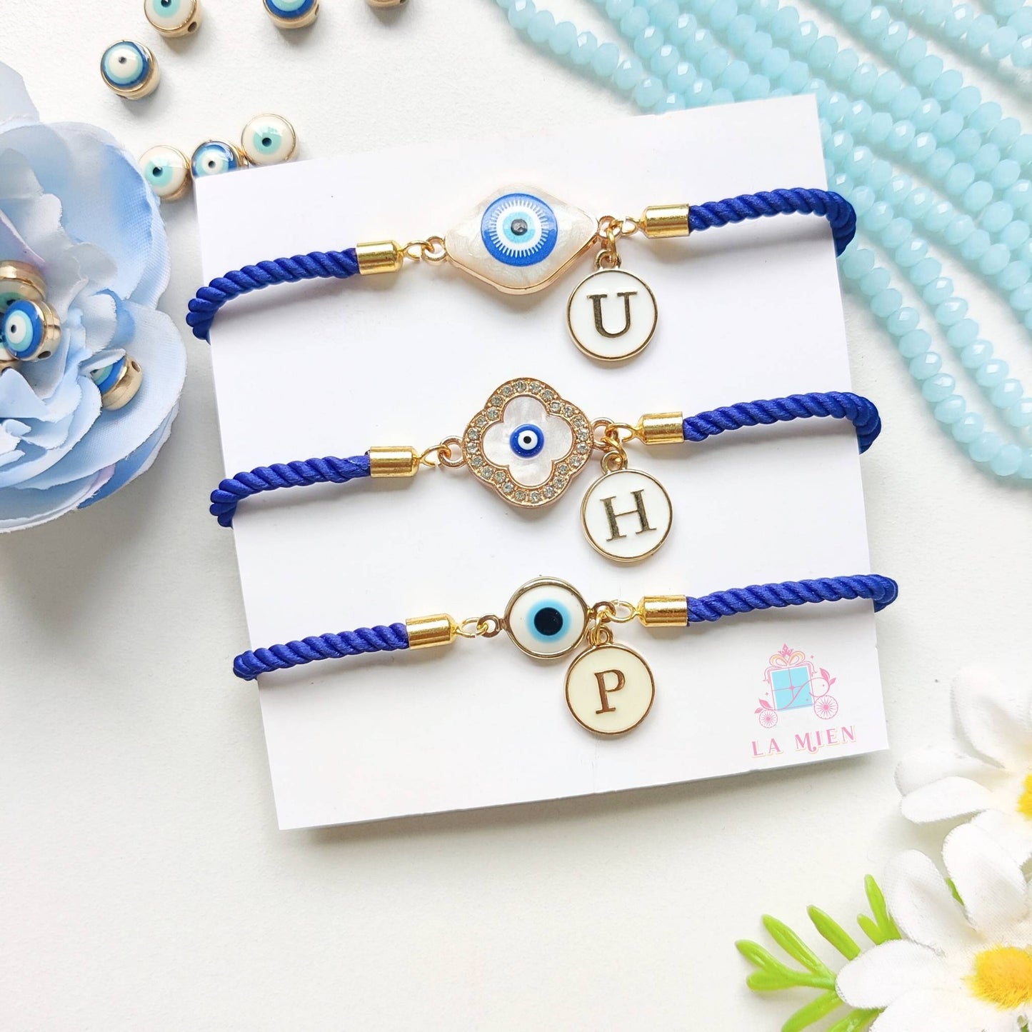 Set of 3 personalized evil eye rakhi bracelets with initial charms and adjustable blue cords.